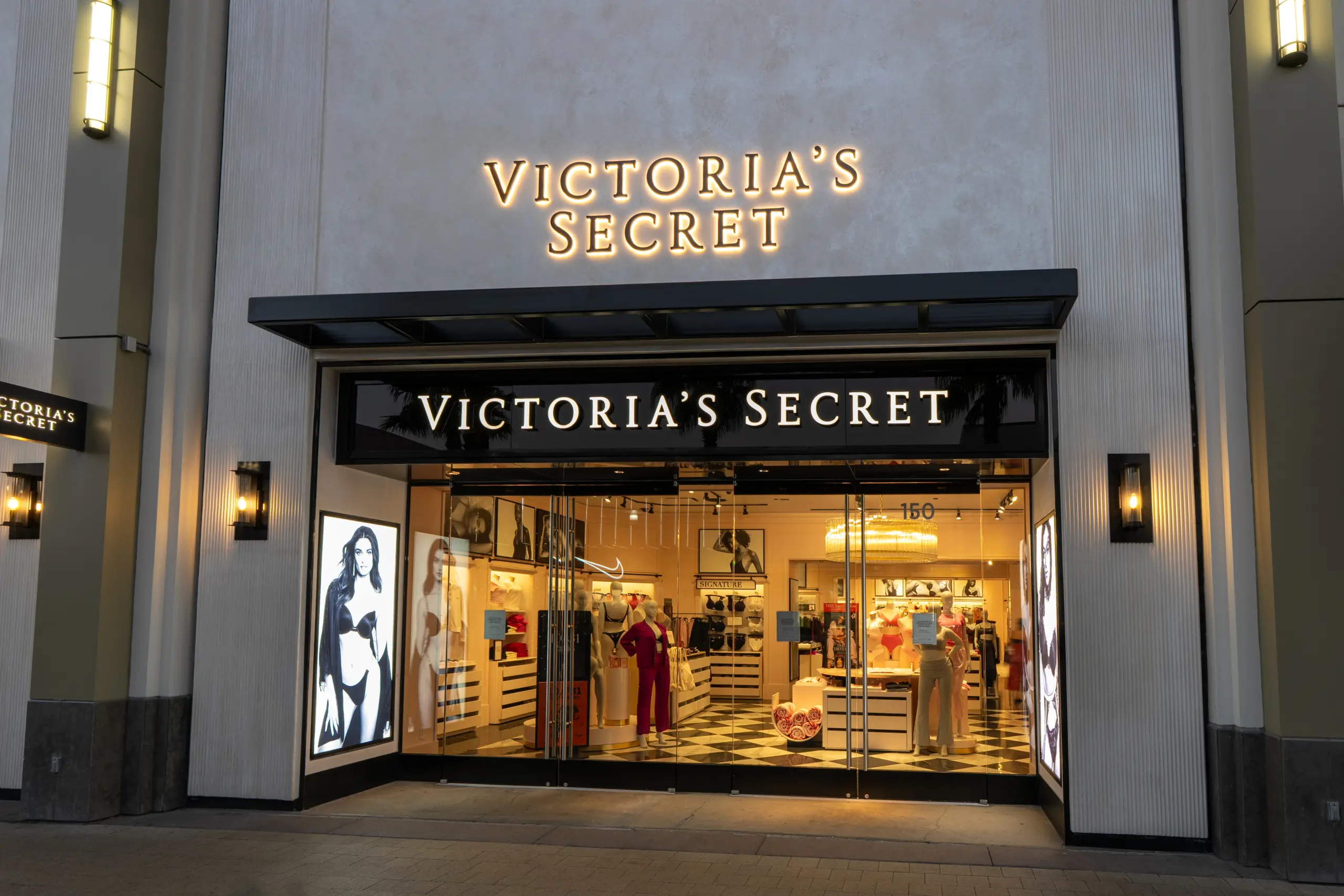 The entrance of a Victoria's Secret store, with illuminated Victoria's Secret signage above the glass doors and displays of lingerie and mannequins visible inside the brightly lit store.