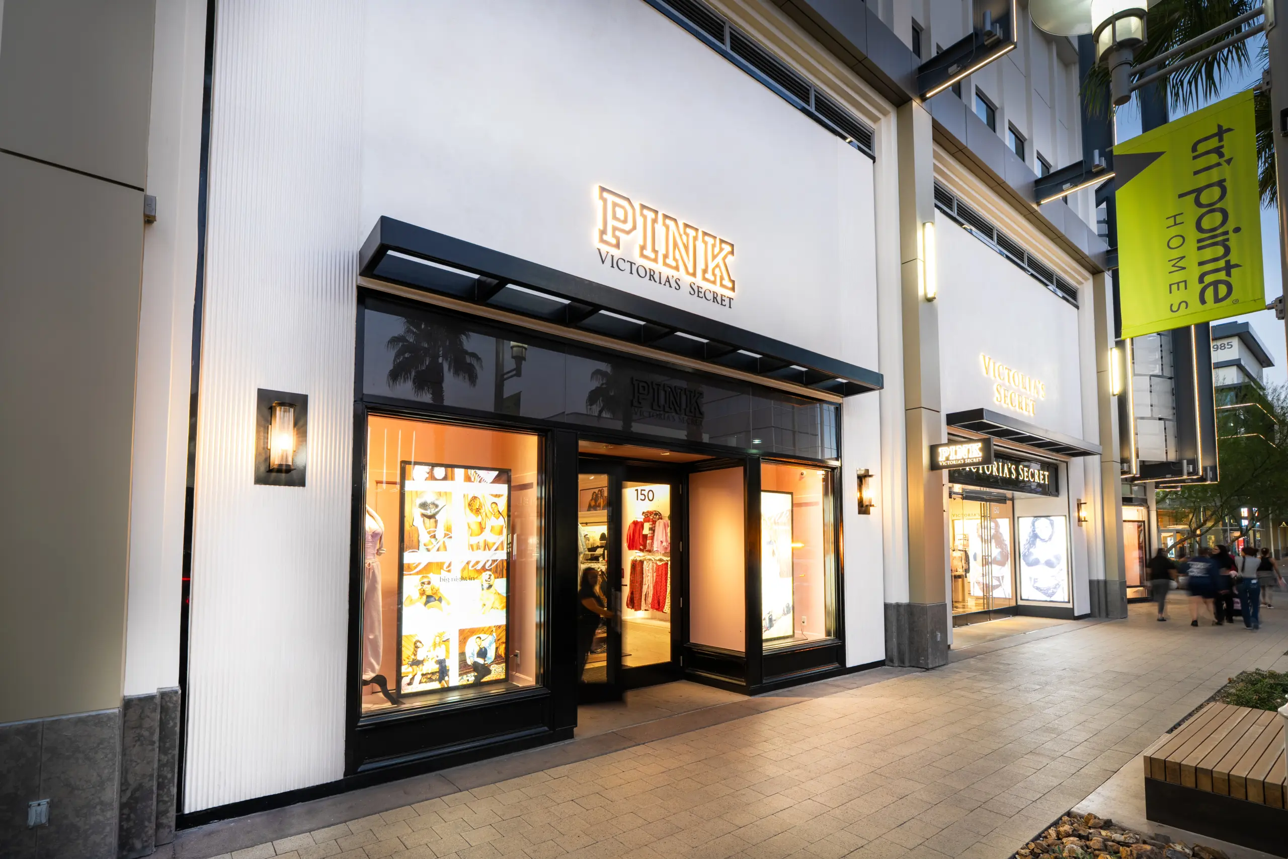 The PINK by Victoria’s Secret storefront at an outdoor shopping center features illuminated window displays of clothing and accessories, with the signature PINK by Victoria Secret branding, neighboring stores, and walkway plants visible.