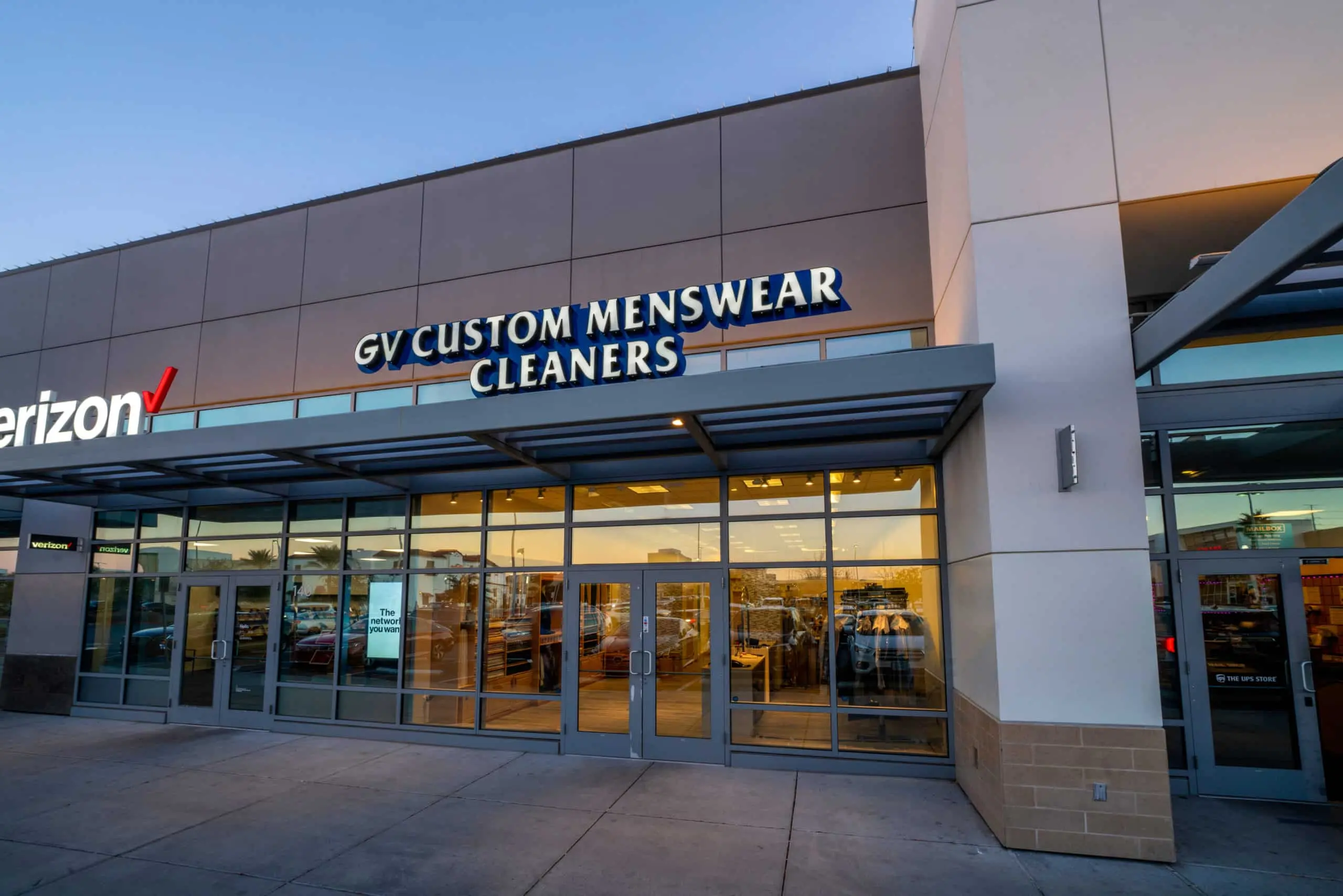 DSC00632-Edit Storefront of GV Custom Menswear Cleaners in a shopping plaza, featuring large glass windows and doors. The neighboring Verizon store sign is partially visible on the left, highlighting the convenient location of GV Custom Menswear.