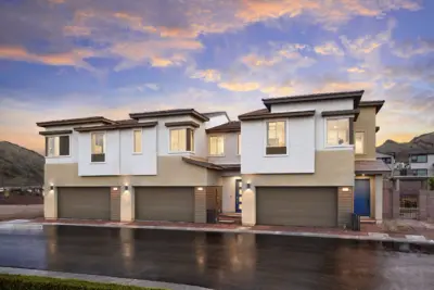 Modern two-story duplex homes with beige and white exteriors, large windows, and three-car garages, photographed at sunset with a mountain backdrop and wet pavement in front.