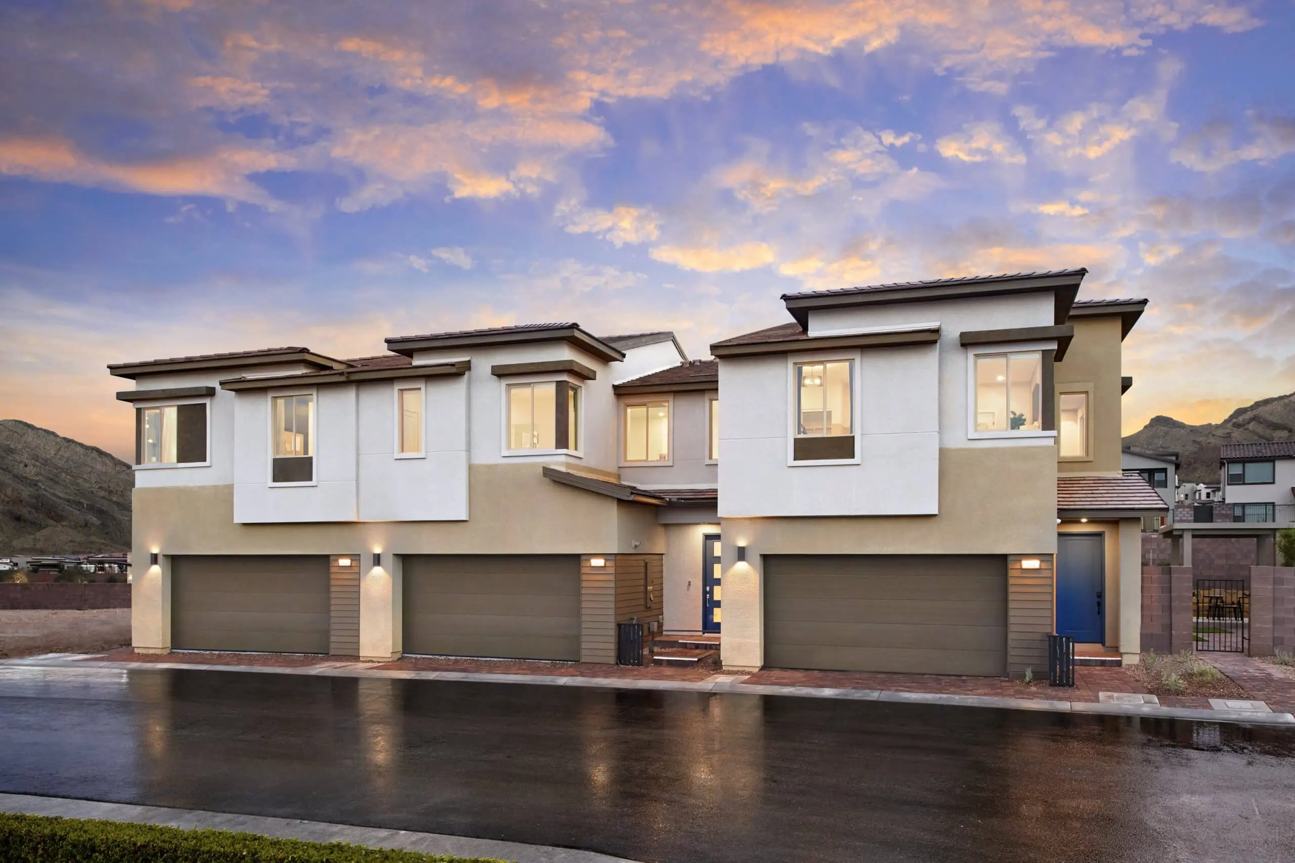 230816_Vertex_Exteriors_001_1 Modern two-story duplex homes with beige and white exteriors, large windows, and three-car garages, photographed at sunset with a mountain backdrop and wet pavement in front.