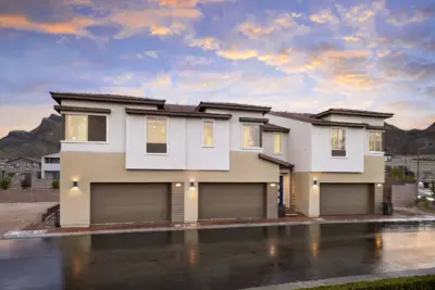 Modern two-story duplex with beige and white exterior, three large garage doors, illuminated lights, and mountain views in the background. The driveway and street appear wet, reflecting the colorful sunset sky.