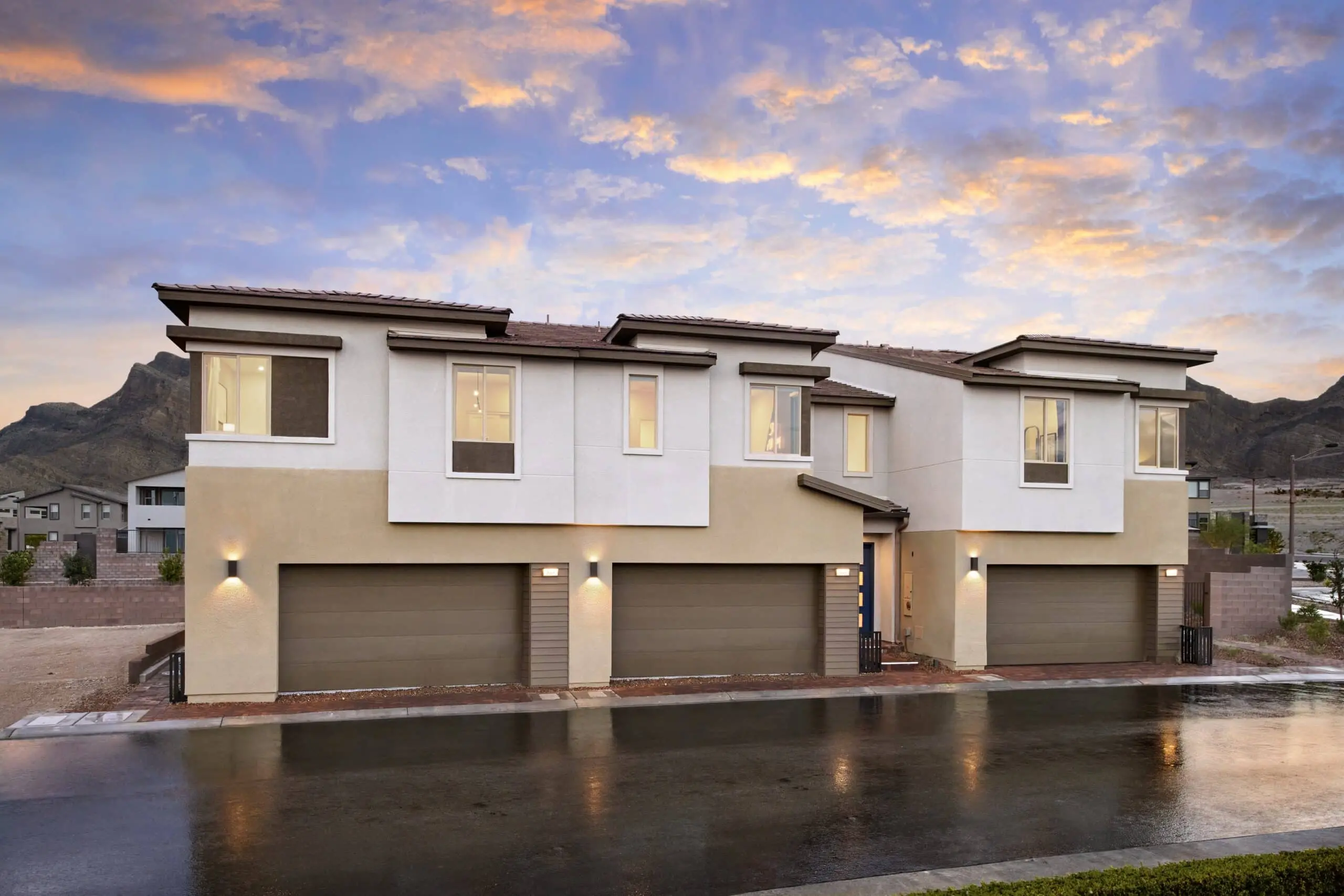 230816_Vertex_Exteriors_002_1 Modern two-story duplex with beige and white exterior, three large garage doors, illuminated lights, and mountain views in the background. The driveway and street appear wet, reflecting the colorful sunset sky.