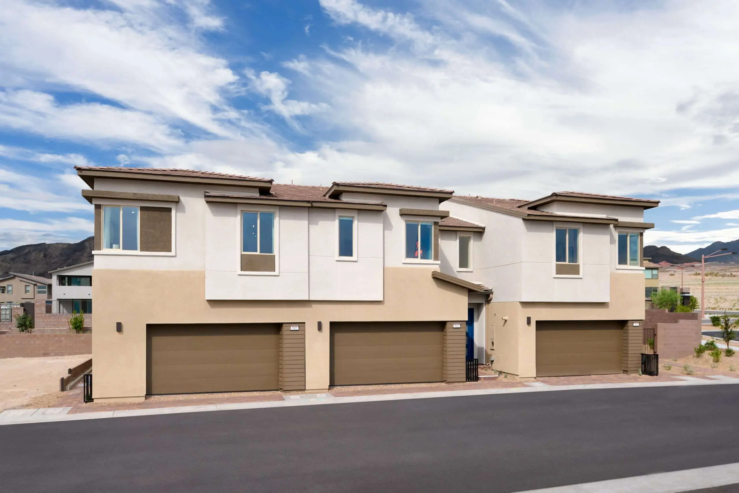 230816_Vertex_Exteriors_003 Two modern beige and white townhouses with three-car garages, flat roofs, and large windows, set against a blue sky with scattered clouds and mountains in the background.