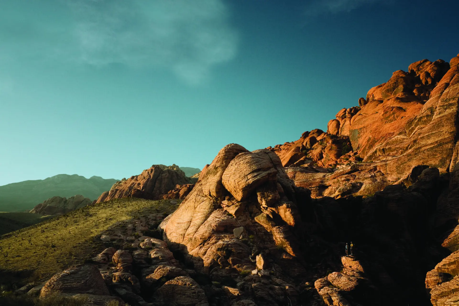 A rocky desert landscape with large red and orange sandstone formations under a blue sky. Sunlight highlights the rock textures, and distant mountains are visible in the background.