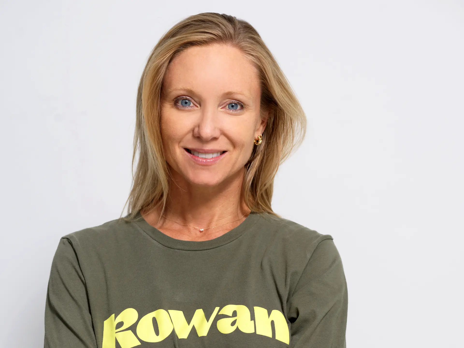 A woman with blonde hair and blue eyes smiles at the camera. She is wearing a green shirt with the word Rowan in yellow letters, and is posed against a plain, light-colored background.