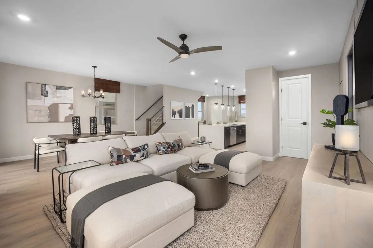 A modern open-concept living space featuring a cozy sectional sofa, round coffee table, wall-mounted TV, dining table with chairs, pendant lights over a kitchen island, and neutral-toned decor with light wood flooring.
