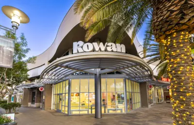 A modern storefront with large glass windows and a prominent Rowan sign is surrounded by palm trees wrapped in string lights, set in a bright outdoor shopping area at dusk.