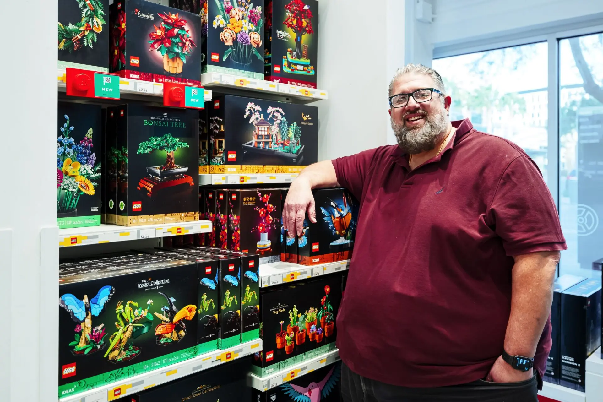 Arthur Rossi Lego master A smiling man in a maroon shirt stands in a store beside shelves filled with LEGO botanical sets, including flowers, a bonsai tree, and butterflies. Bright daylight streams in from windows in the background.
