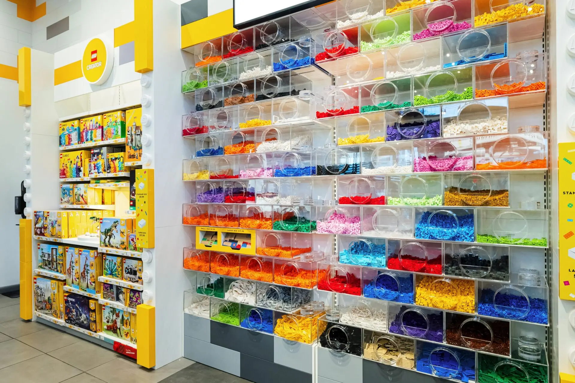 Arthur Rossi Lego master Wall of clear bins filled with LEGO bricks sorted by color, including red, orange, yellow, green, blue, pink, and more. LEGO sets are displayed on shelves to the left in a brightly lit store.