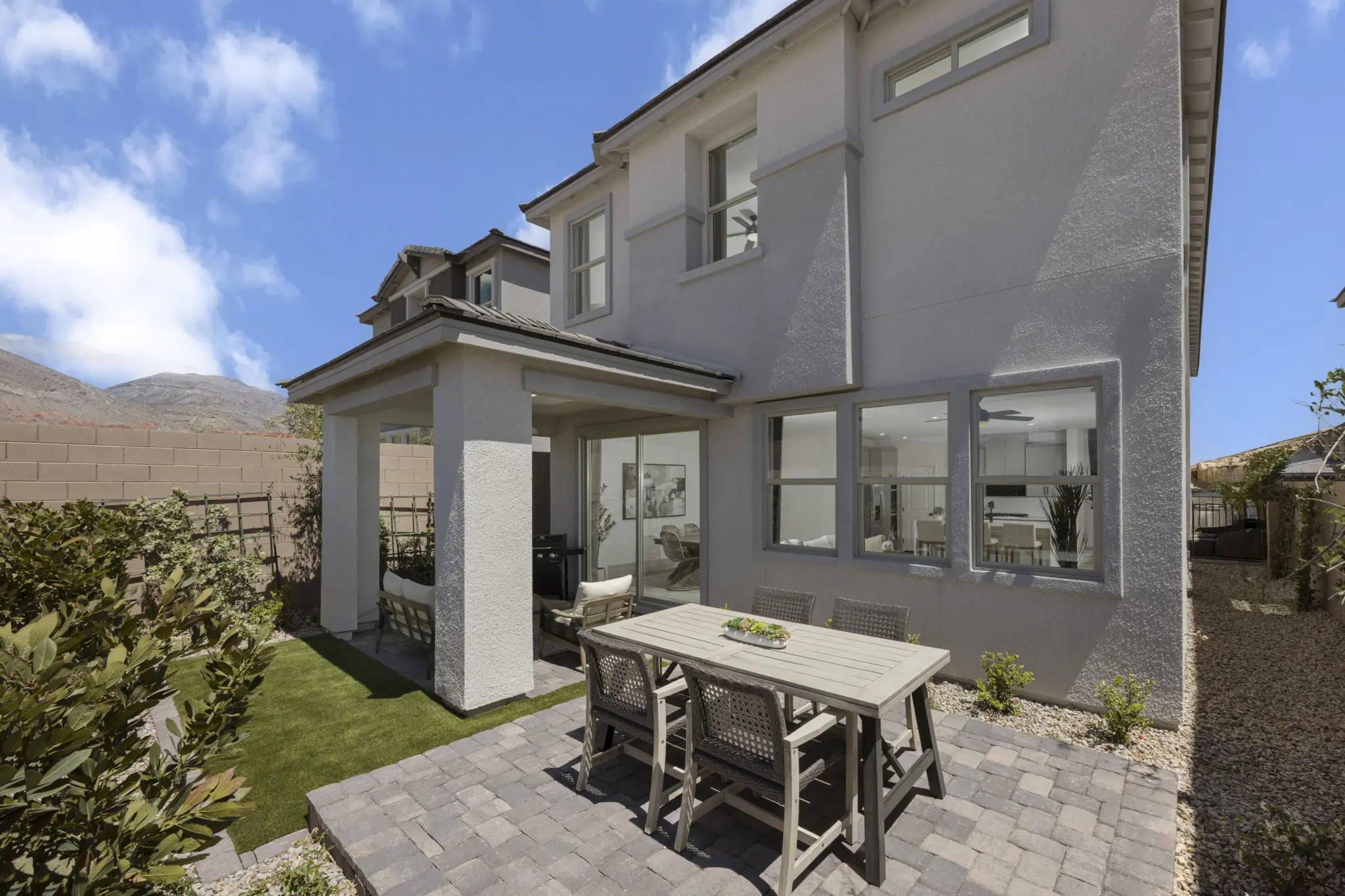 KBLV_LandingsAtCaldwellPark_Plan2070-RearExterior_7250a-11×8-1-2048×1365 A modern two-story house with a light gray exterior, large windows, and a covered patio. Plan 2070 at Caldwell Park by KB Home features an outdoor dining area on stone paving, surrounded by lush plants and a grass lawn under a blue sky.