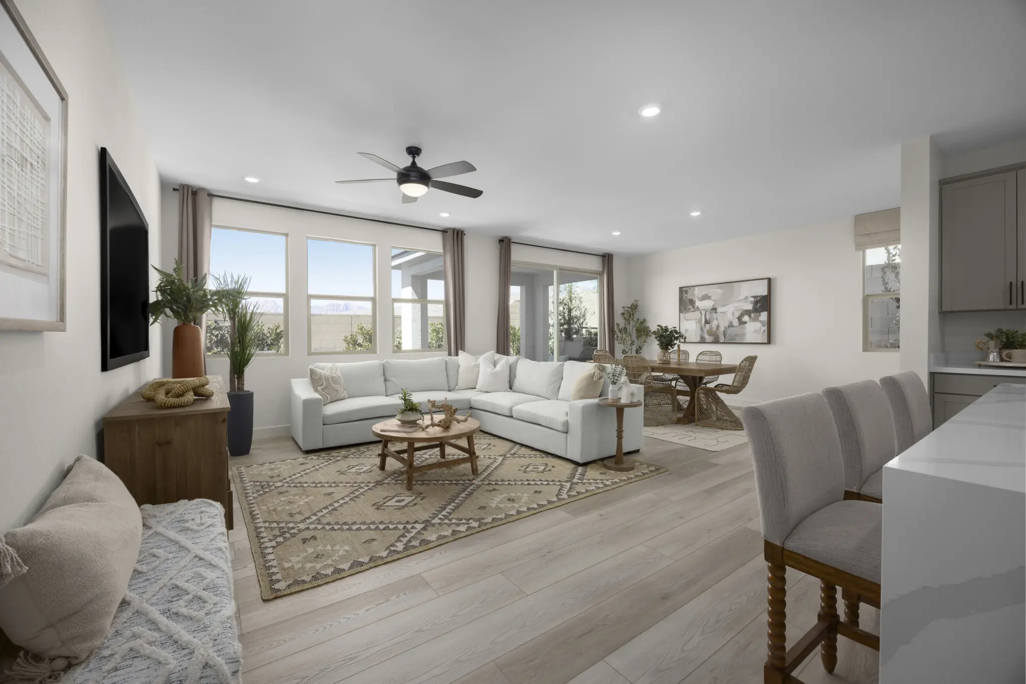 KBLV_LandingsatCaldwellPark_Plan2070-Greatroom_7163b-11×8-1-2048×1365 Bright, modern living and dining area in Plan 2070 at Caldwell Park by KB Home with light wood floors, a white sectional sofa, patterned rug, round coffee table, large windows, indoor plants, and a dining table near the open kitchen island with barstools.
