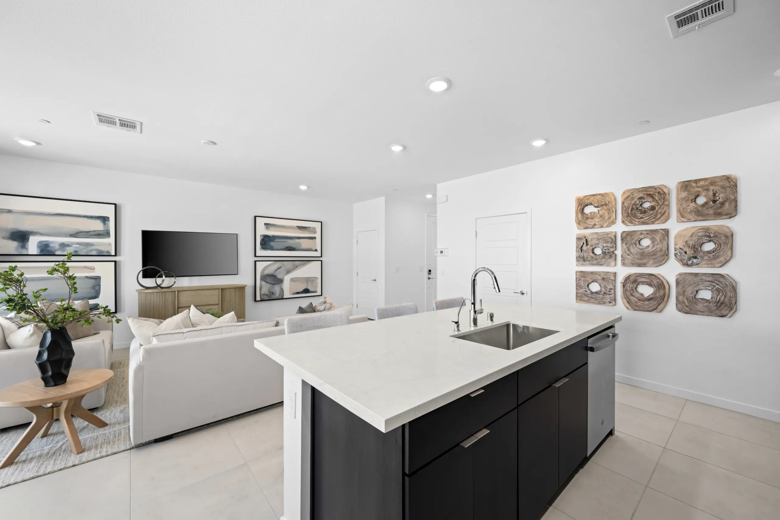 NEV_Sandpiper_Summerlin_Pic_Jan_Kitchen_3of5_F1 Modern open-concept living room and kitchen in the Jan Model at Sandpiper by Lennar, featuring white walls, a central island with sink, light sofas, abstract wall art, TV, and decorative wood pieces. Bright, minimalist decor.