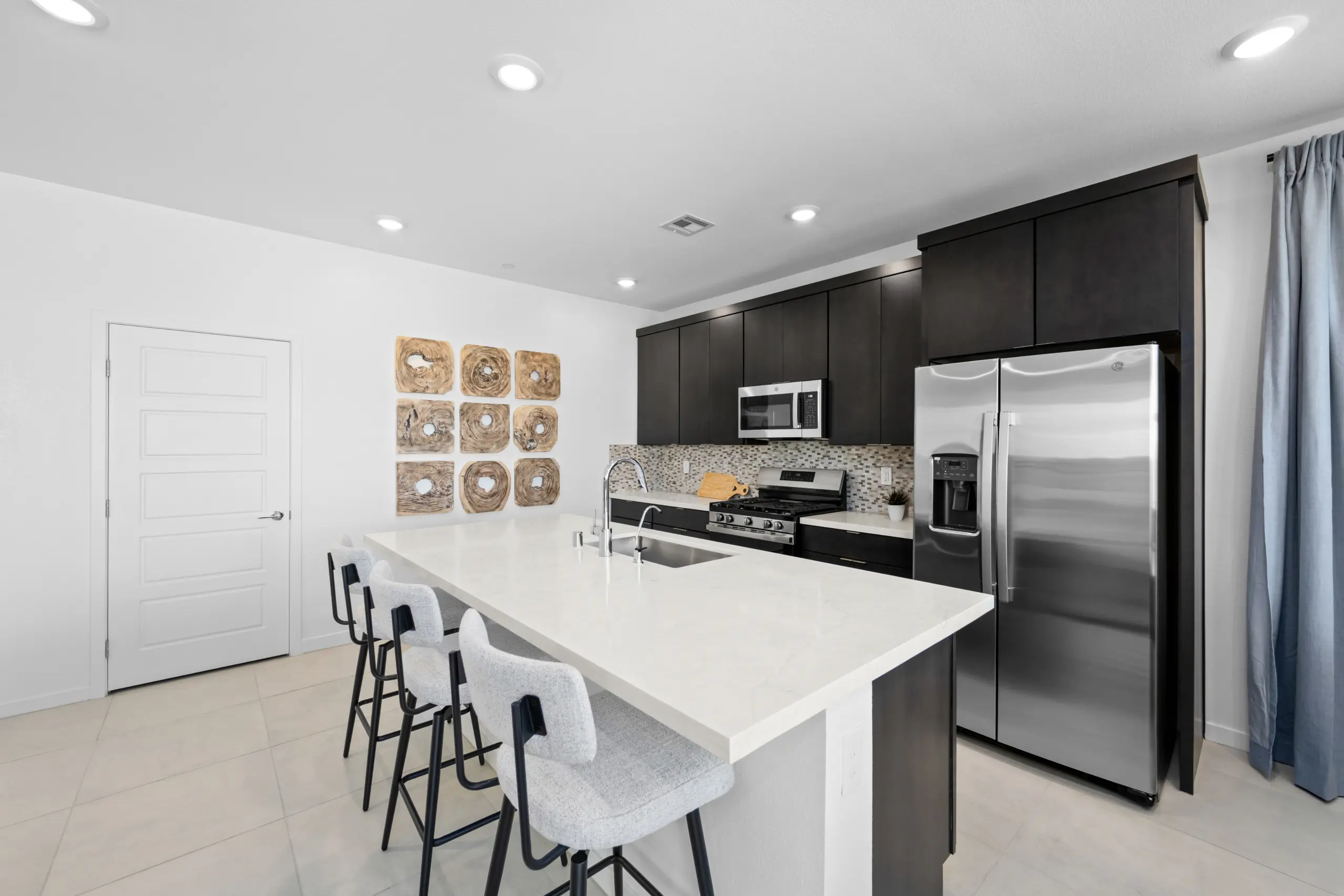 NEV_Sandpiper_Summerlin_Pic_Jan_Kitchen_4of5_F1 Modern kitchen in the Jan Model at Sandpiper by Lennar features dark cabinets, stainless steel appliances, a white island with sink, three gray bar stools, decorative wall art, blue curtains by the refrigerator, and a light tile floor.