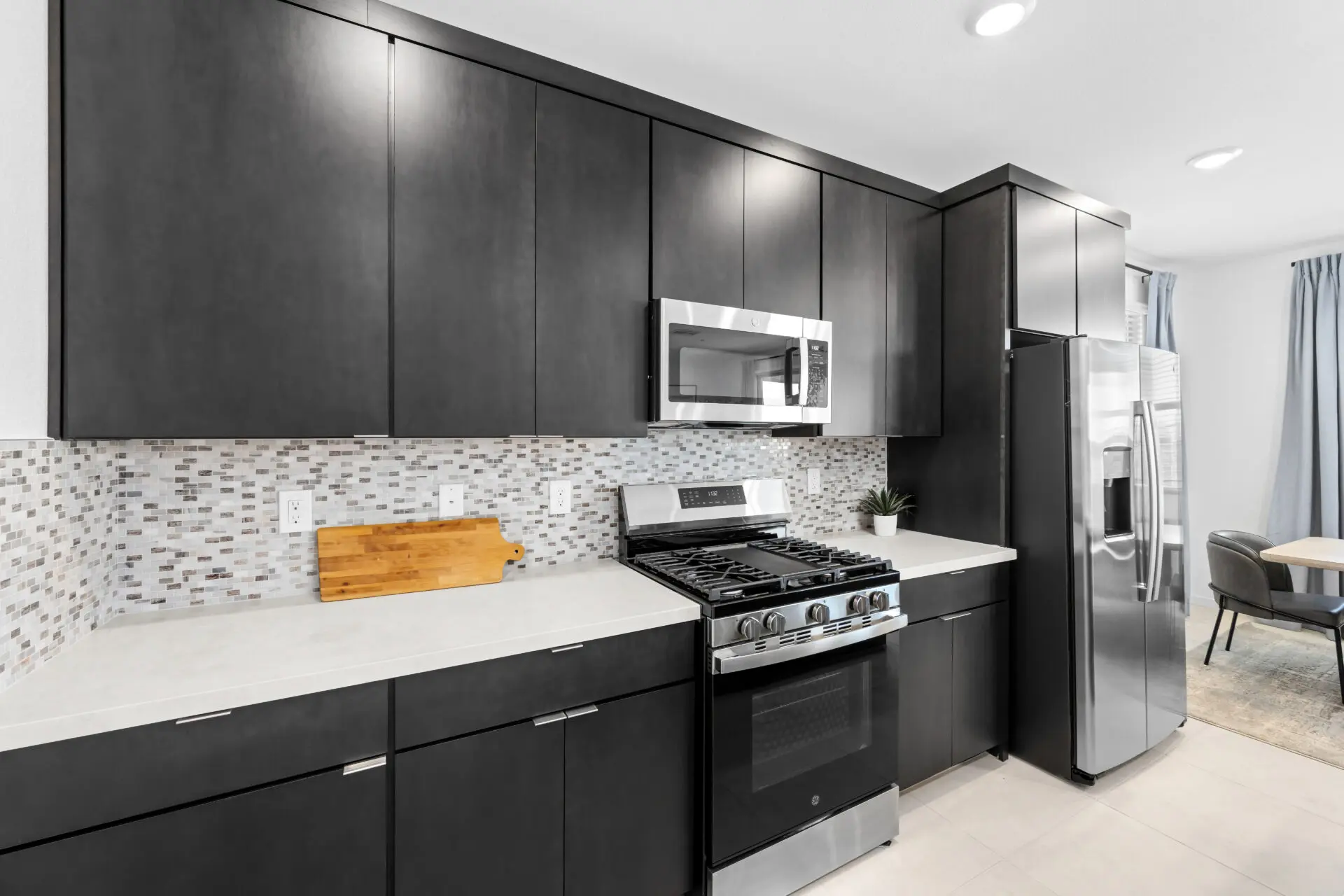 NEV_Sandpiper_Summerlin_Pic_Jan_Kitchen_5of5_F1 Modern kitchen in the Jan Model at Sandpiper by Lennar features dark cabinets, stainless steel appliances, a mosaic tile backsplash, white countertops, a small cutting board, and a potted plant for a stylish touch.