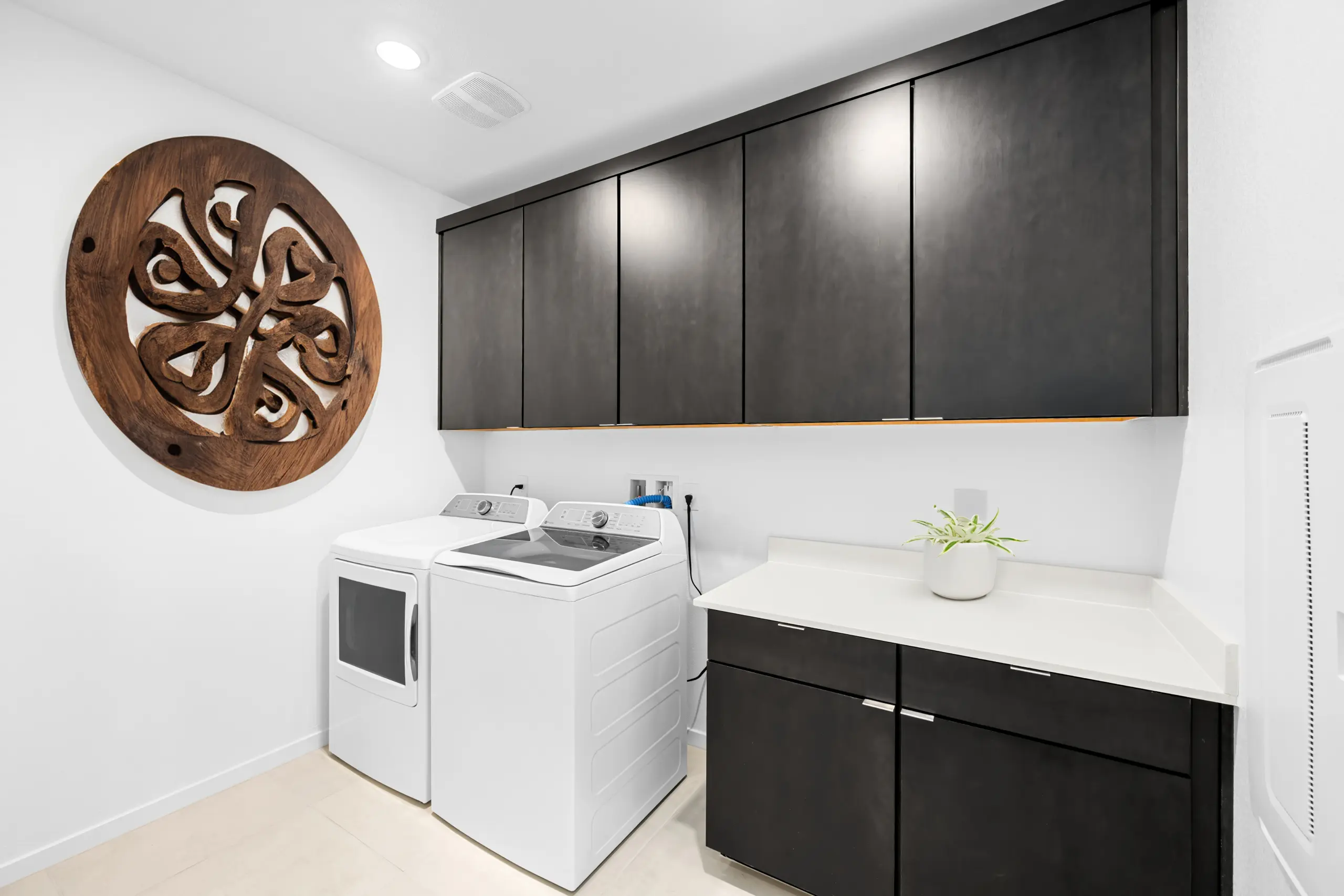 NEV_Sandpiper_Summerlin_Pic_Jan_Laundry_F2 Modern laundry room in the Jan Model at Sandpiper by Lennar, featuring a white washer and dryer, dark cabinets above and below a white countertop, a small potted plant, and large decorative wooden wall art on the left wall.