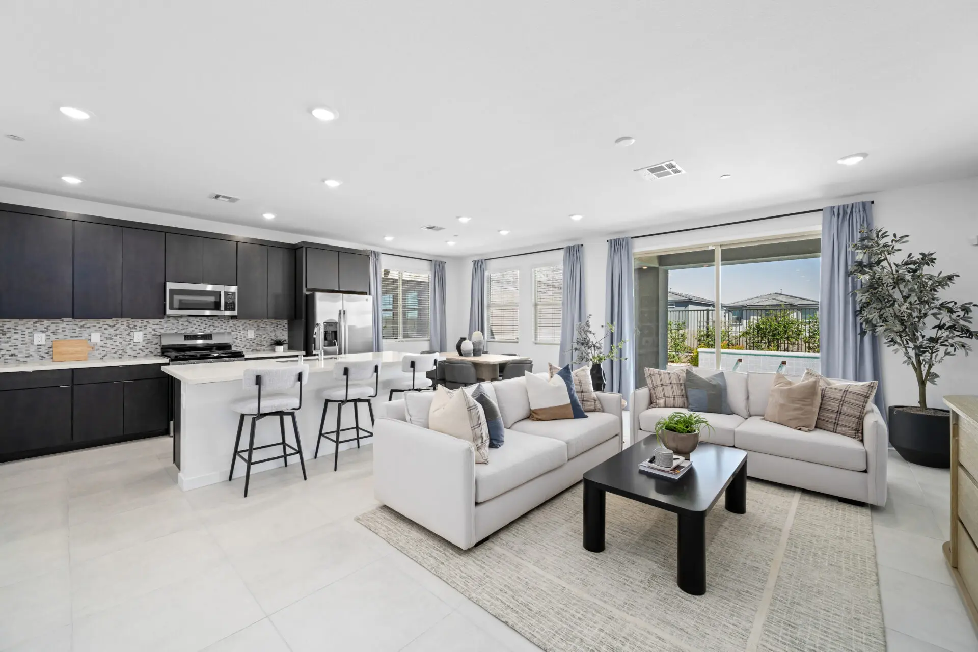 NEV_Sandpiper_Summerlin_Pic_Jan_Living_1of4_F1 Experience the Jan Model at Sandpiper by Lennar—an open-concept living space with a modern kitchen featuring dark cabinets, an island with bar stools, and a bright living area with white sofas and large windows.