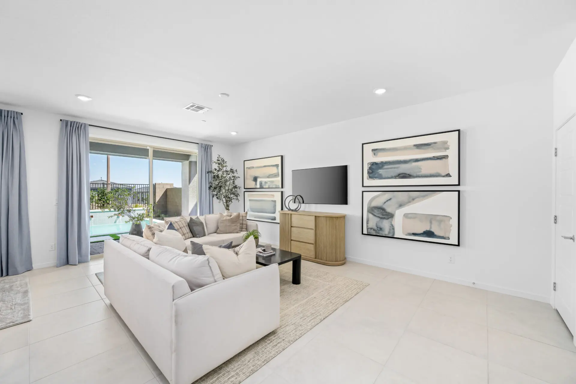 NEV_Sandpiper_Summerlin_Pic_Jan_Living_2of4_F1 Bright, modern living room in the Jan Model at Sandpiper by Lennar, featuring a white sofa, light tile flooring, abstract artwork, TV on a wooden console, large windows with blue curtains, and a view of the patio and pool.