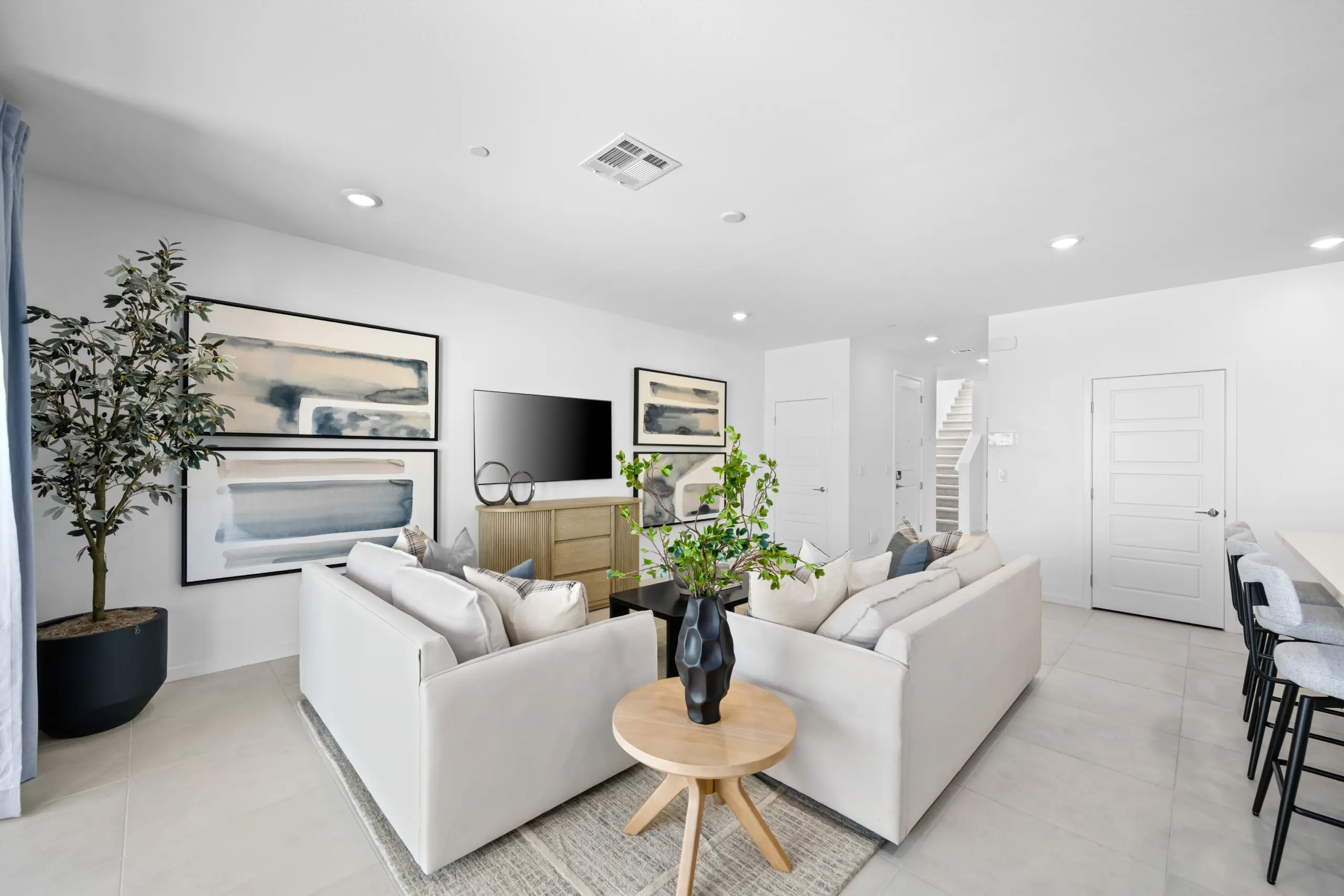 NEV_Sandpiper_Summerlin_Pic_Jan_Living_3of4_F1 Modern living room in the Jan Model at Sandpiper by Lennar features two light-colored sofas, a round wooden table with a plant, abstract wall art, a TV console, tall potted plant, and kitchen bar with stools in a bright, open space.