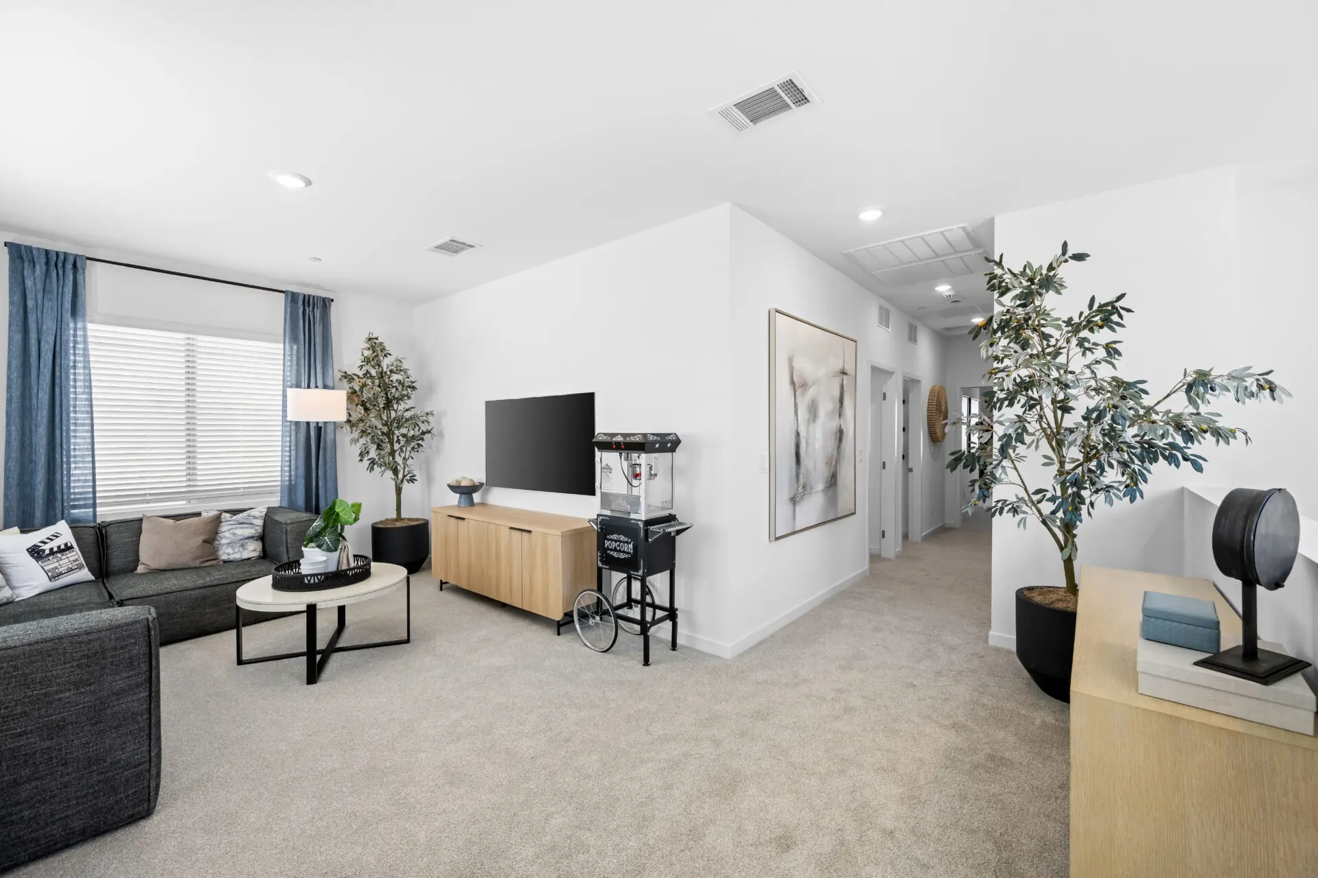 NEV_Sandpiper_Summerlin_Pic_Jan_Loft_1of3_F2 Modern living room in the Jan Model at Sandpiper by Lennar, featuring a gray sectional sofa, round coffee table, large TV on a light wood stand, popcorn machine, potted plants, and bright neutral tones filled with natural light.