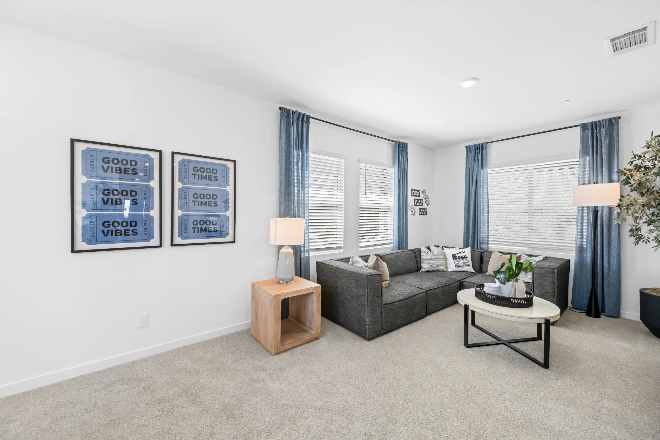 NEV_Sandpiper_Summerlin_Pic_Jan_Loft_2of3_F2 A modern living room in the Jan Model at Sandpiper by Lennar features a gray sectional sofa, blue curtains, a round coffee table with plants, framed GOOD VIBES and GOOD TIMES art on the wall, and large windows letting in natural light.
