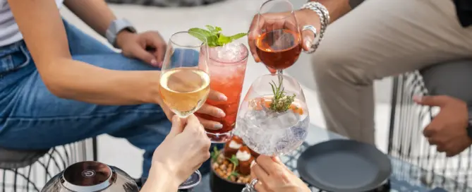Four people making a toast with various colorful cocktails and wine glasses at an outdoor table, with snacks and empty plates in the background.