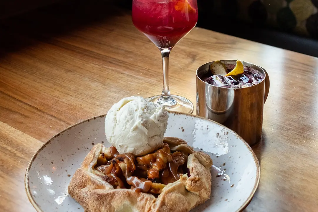 A rustic apple tart topped with vanilla ice cream sits on a plate beside a red cocktail in a wine glass and a dark drink in a metal cup, all on a wooden table.