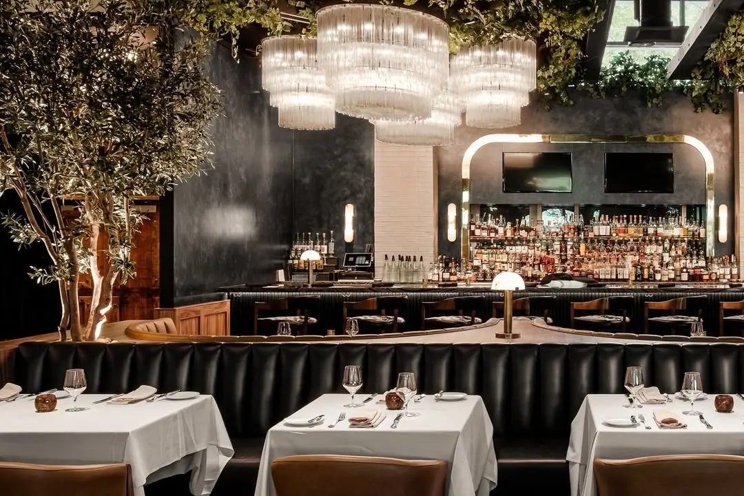 A stylish restaurant interior with black leather booths, white tablecloths, elegant place settings, a well-stocked bar, large mirrors, and modern chandeliers hanging from a ceiling decorated with greenery.