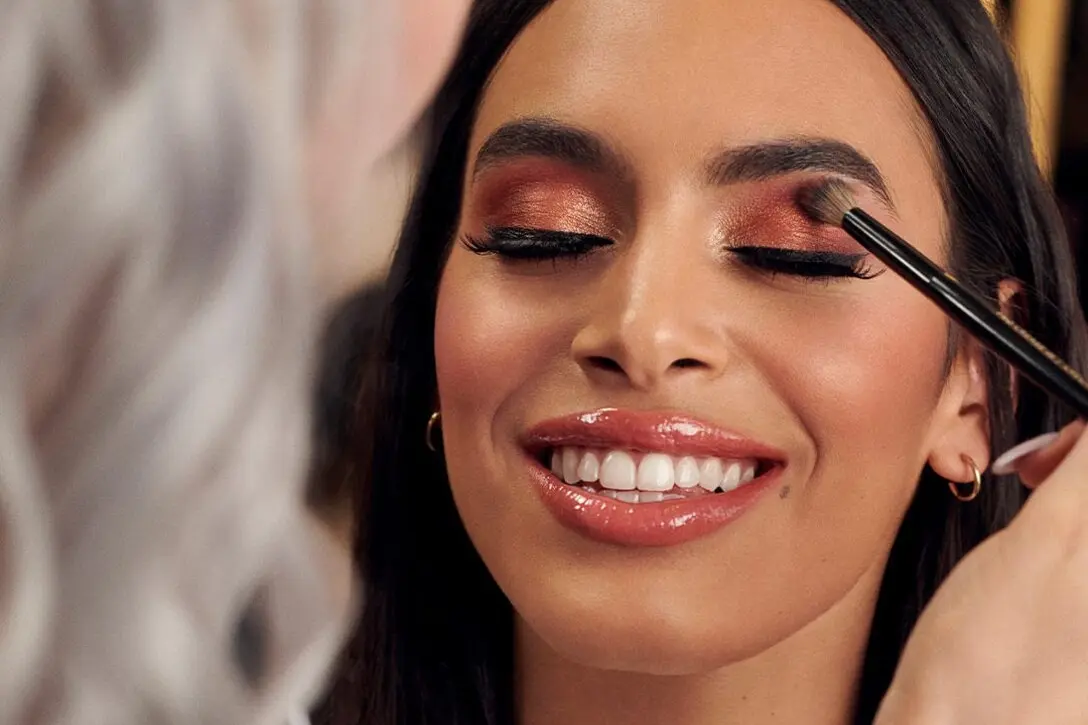 A smiling woman with shiny lip gloss and metallic copper eyeshadow is having her eye makeup applied with a brush by another person. Her eyes are closed and she appears relaxed.