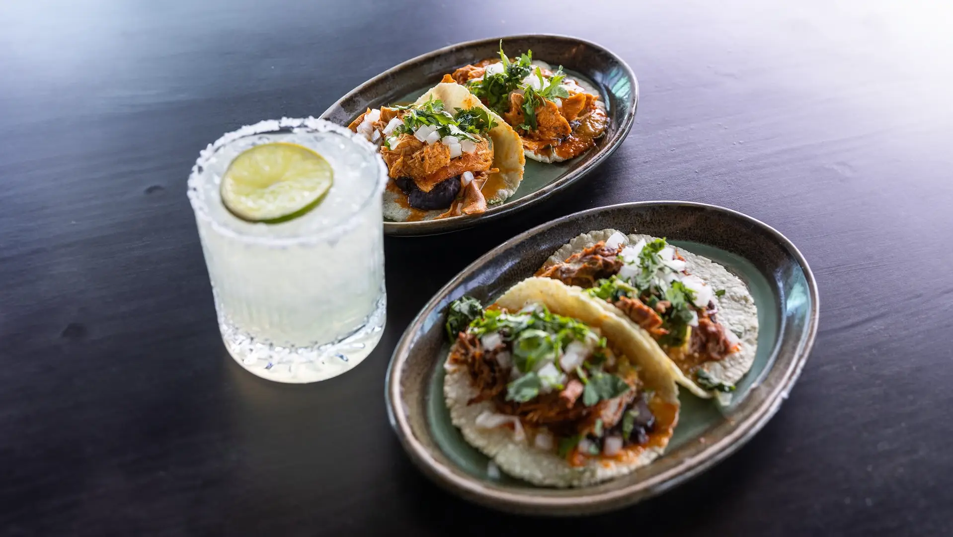Two plates, each with two tacos filled with meat, onions, and cilantro, sit on a dark table next to a glass of margarita garnished with a salted rim and a slice of lime.