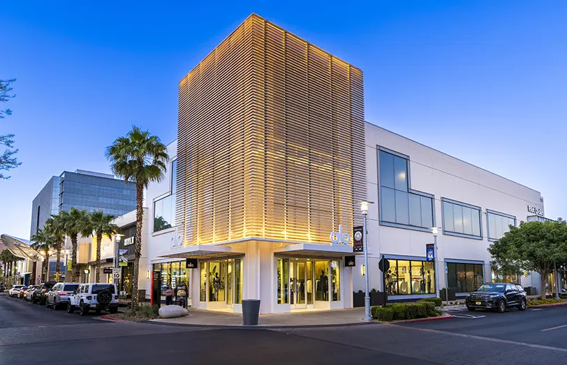 Alo 2026 (1) A modern two-story building with large windows and a striking illuminated lattice structure at the corner, surrounded by palm trees, parked cars, and clear evening skies.