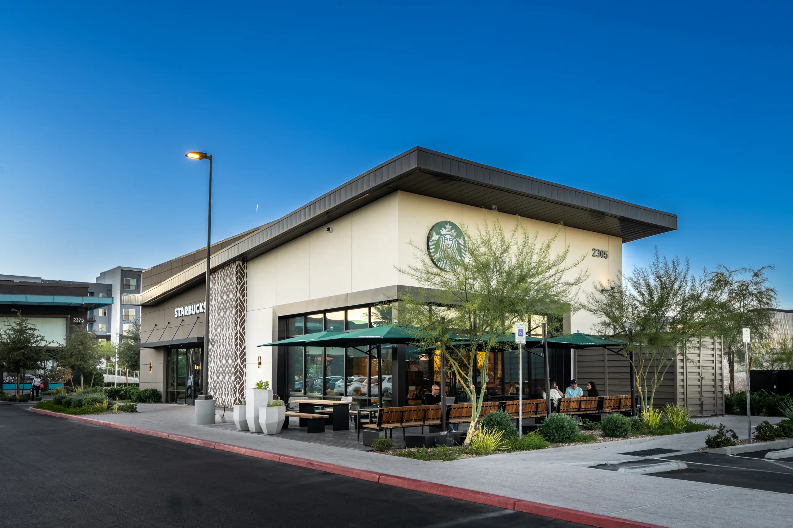 A modern Starbucks café with outdoor seating, green umbrellas, and large windows sits on a landscaped corner under a clear blue sky. A few people are seated outside, and trees are planted around the building.