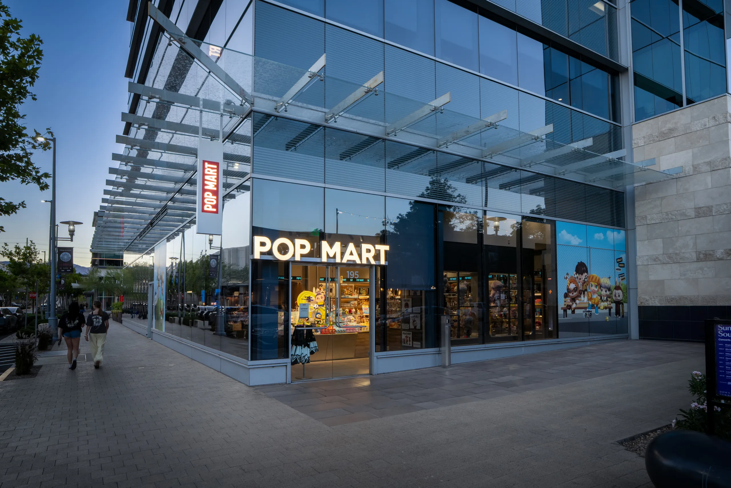 A modern POP MART store with large glass windows and bright signage is on a city sidewalk. Colorful toy displays and cartoon graphics are visible inside and on the windows. People walk by under a glass awning.