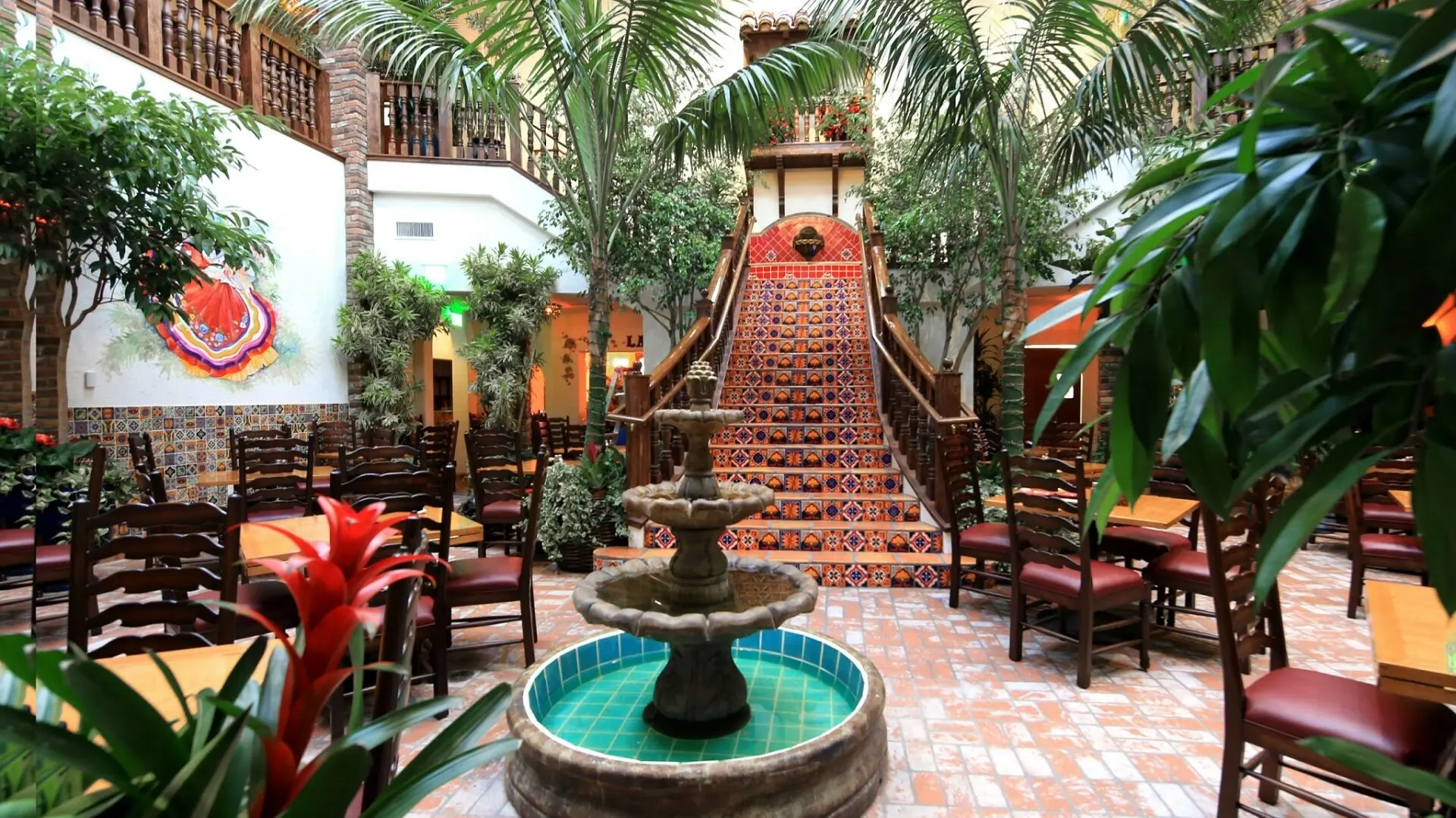 A vibrant indoor courtyard with palm trees, colorful tiled steps, a central fountain, wooden chairs, and lush greenery, creating a warm, inviting Mexican or Spanish-style atmosphere.