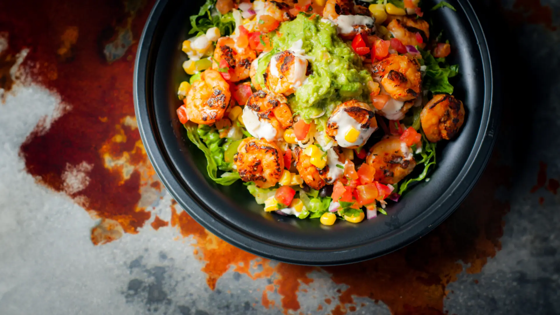 A black bowl filled with grilled shrimp, guacamole, diced tomatoes, corn, onions, and lettuce, sitting on a rustic, weathered surface.