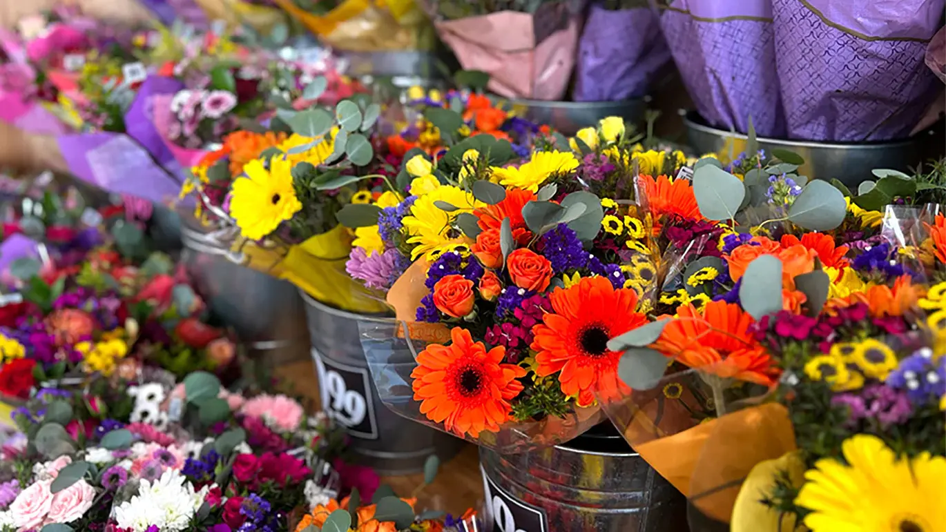 Buckets filled with vibrant, mixed flower bouquets—including orange gerbera daisies, yellow chrysanthemums, purple statice, and greenery—are displayed for sale, wrapped in colorful paper on a market stall.