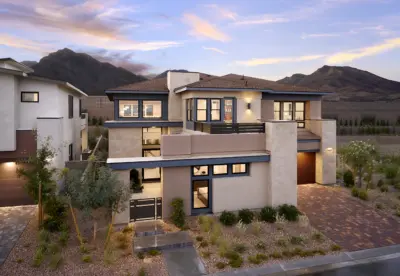 Modern two-story house with large windows, a flat roof, and a mix of stone and stucco exterior—Plan 3 at Carlisle Peak by Tri Pointe Homes—surrounded by desert landscaping and mountains in the background at sunset.