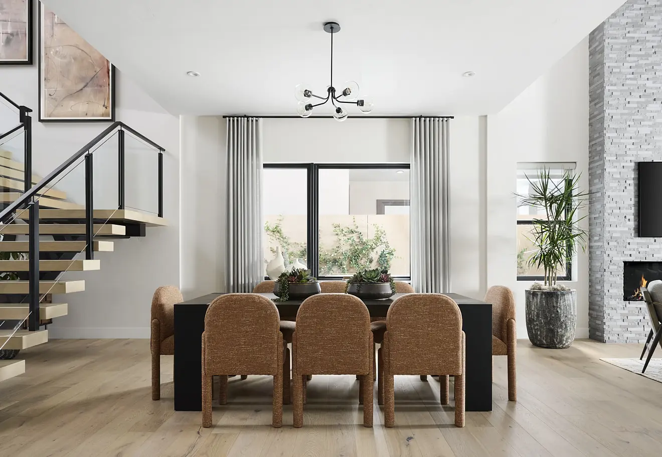 250911_Carlisle_Peak_P3_004 Modern dining room in Plan 3 at Carlisle Peak by Tri Pointe Homes, featuring a black rectangular table, eight tan upholstered chairs, large window with white curtains, indoor plants, light wood floors, and a floating staircase with glass railing.