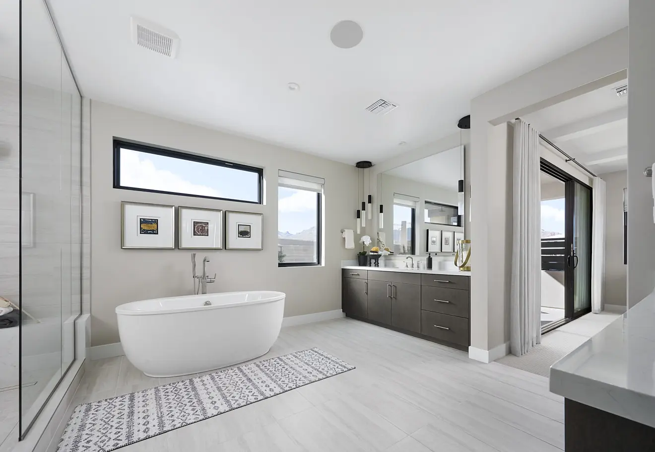 250911_Carlisle_Peak_P3_007 Modern bathroom in Plan 3 at Carlisle Peak by Tri Pointe Homes features a freestanding white bathtub, large double-sink vanity, wall-mounted mirrors, glass shower, framed art, patterned rug, and bright natural light from windows and a balcony door.