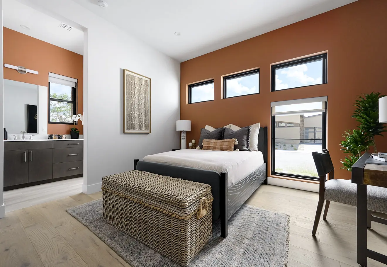 250911_Carlisle_Peak_P3_013 Modern bedroom in Plan 3 at Carlisle Peak by Tri Pointe Homes features an orange accent wall, three horizontal windows, neutral bedding, wicker storage bench, desk with chair, potted plant, and ensuite bath with dark cabinets and white countertops.