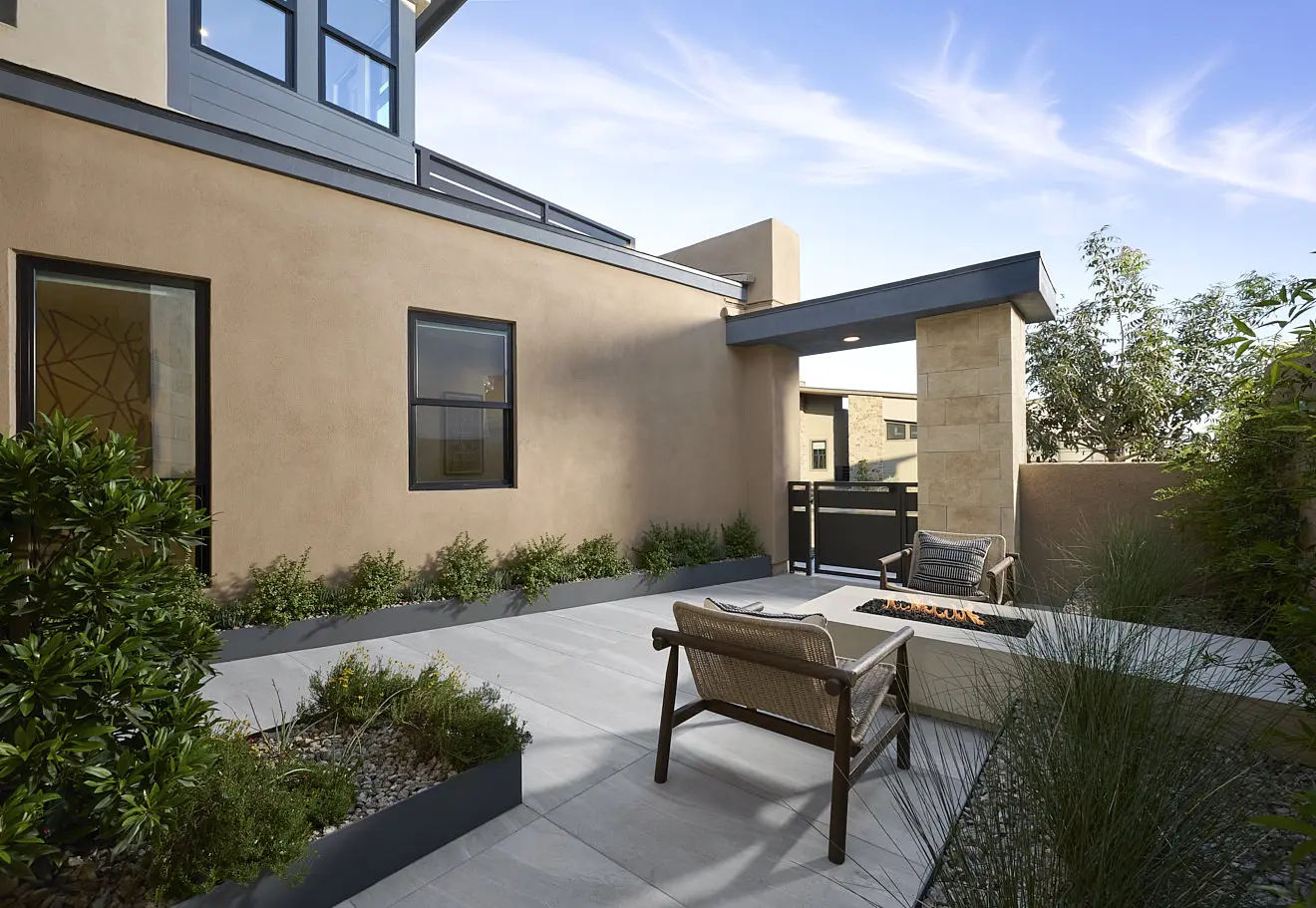 250911_Carlisle_Peak_P3_015 A modern outdoor patio at Plan 3 at Carlisle Peak by Tri Pointe Homes features a fire pit, two wooden chairs with cushions, potted plants, and light stone tile flooring, surrounded by beige walls and greenery under a clear blue sky.