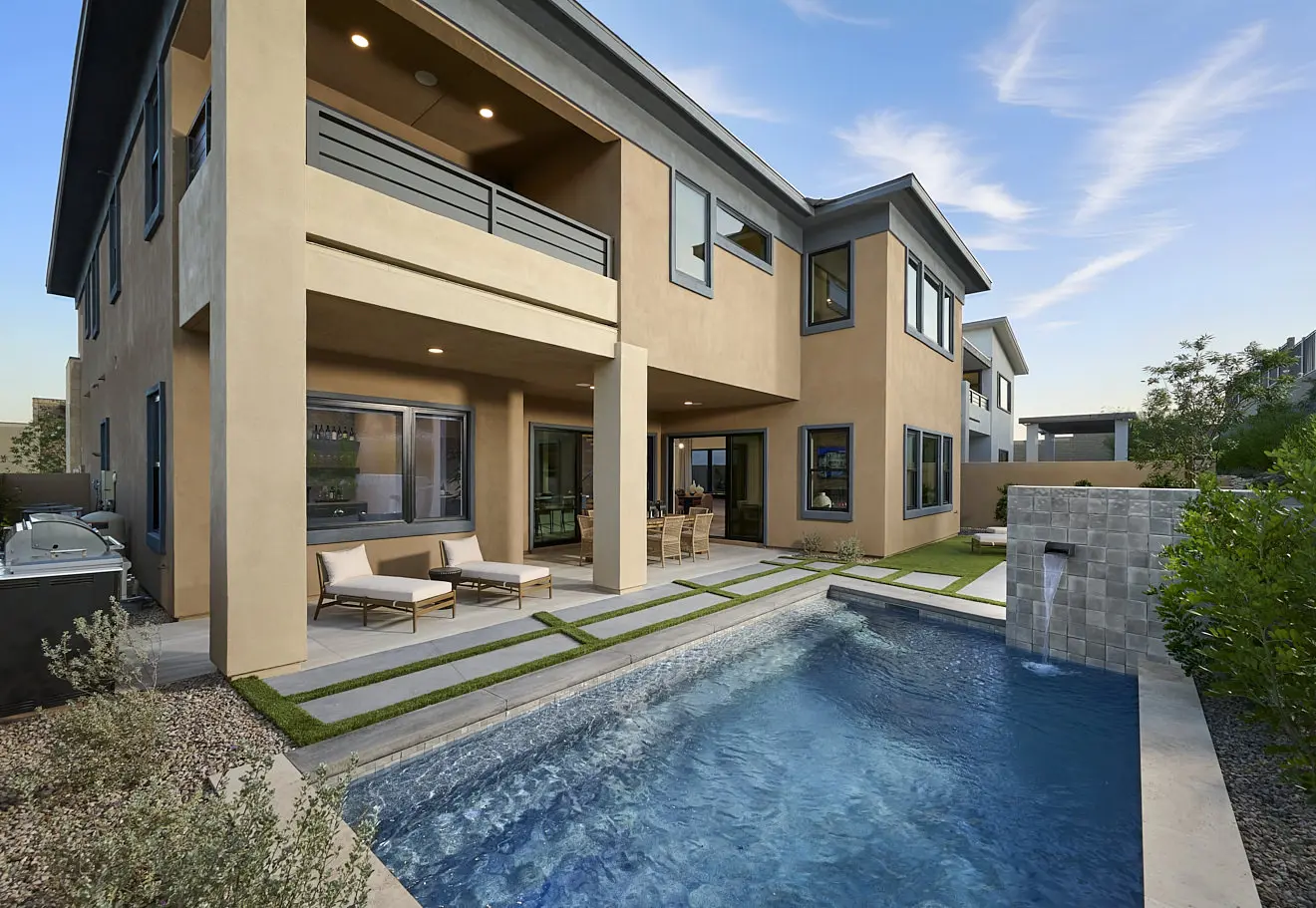 250911_Carlisle_Peak_P3_016 Modern two-story house with tan walls, large windows, and two balconies. The backyard at Plan 3 at Carlisle Peak by Tri Pointe Homes features a small rectangular pool, patio seating, a dining area, and landscaping with grass and stone paths.