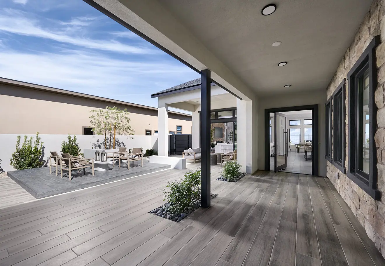 250911_Carlisle_Ridge_P1_002 Modern outdoor patio at Plan 1 at Carlisle Ridge by Tri Pointe Homes features wood-like flooring, a covered walkway, and seating with tables and chairs. Stone wall accents, large windows, blue sky, and lush landscaping complete the scene.