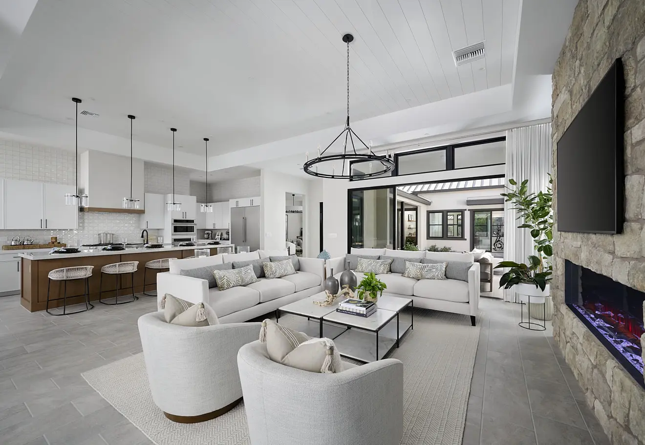 250911_Carlisle_Ridge_P1_003 Modern open-concept living at Plan 1 at Carlisle Ridge by Tri Pointe Homes, featuring a kitchen and living room with white sofas, armchairs, a stone fireplace with electric fire, pendant lighting, large windows to the patio, plants, and neutral accents.