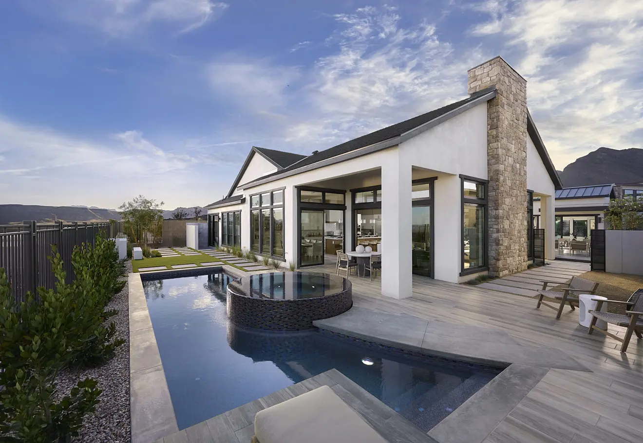 250911_Carlisle_Ridge_P1_005 Modern house with large glass windows, a stone chimney, and an outdoor area featuring a pool, spa, and lounge spaces. Mountains and a blue sky provide the perfect backdrop for Plan 1 at Carlisle Ridge by Tri Pointe Homes.