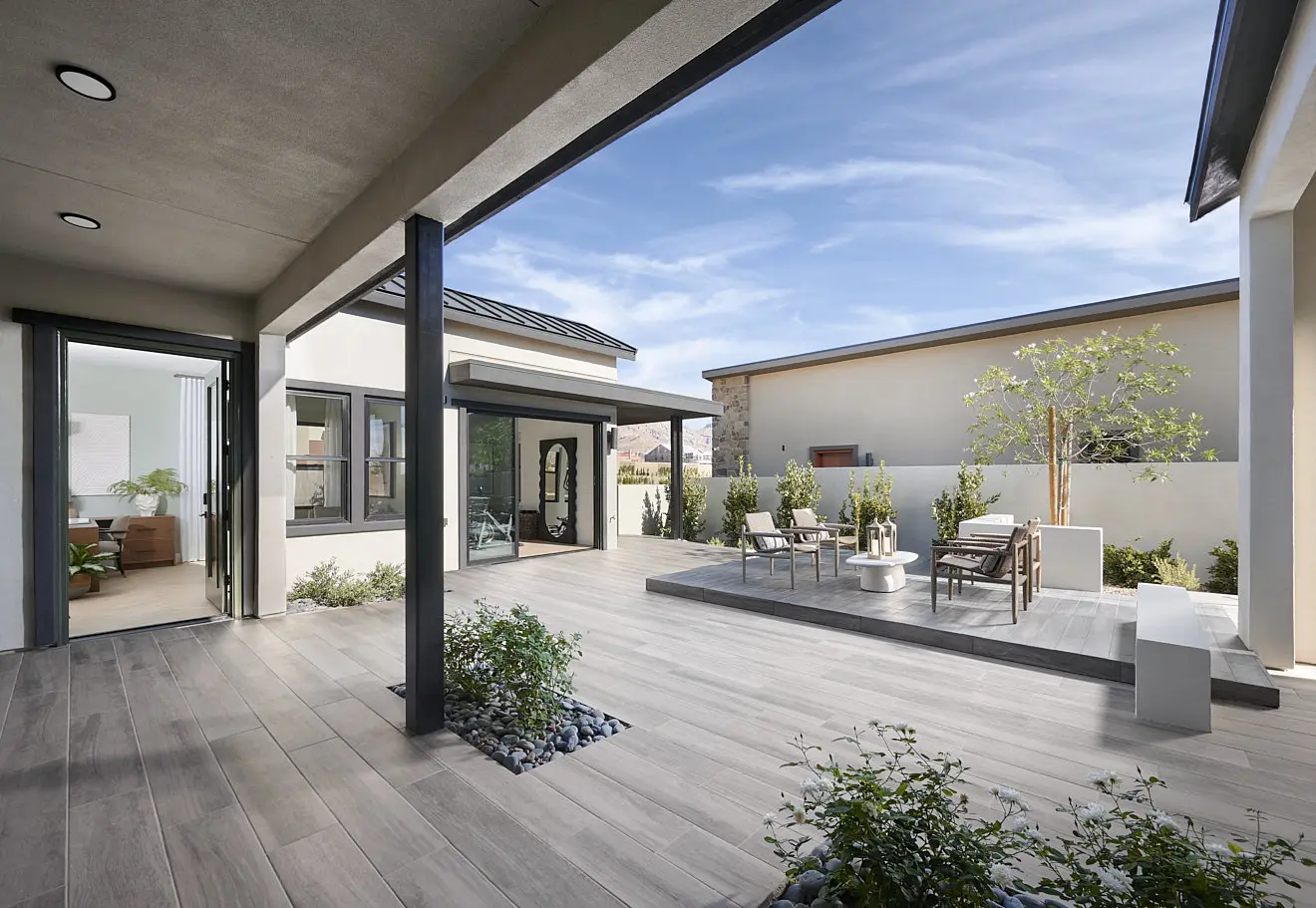 250911_Carlisle_Ridge_P1_006 Modern outdoor patio with light wood flooring, a raised seating area with chairs and a table, potted plants, and large windows; surrounded by white walls and blue sky—an inviting space found in Plan 1 at Carlisle Ridge by Tri Pointe Homes.