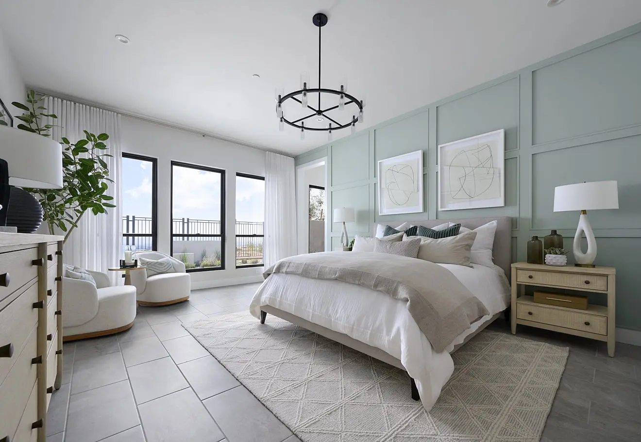 250911_Carlisle_Ridge_P1_008 Modern bedroom in Plan 1 at Carlisle Ridge by Tri Pointe Homes features a light green accent wall, large bed with white bedding, nightstands with lamps, two armchairs by tall windows, dresser, potted plant, and geometric wall art. Natural light fills the room.