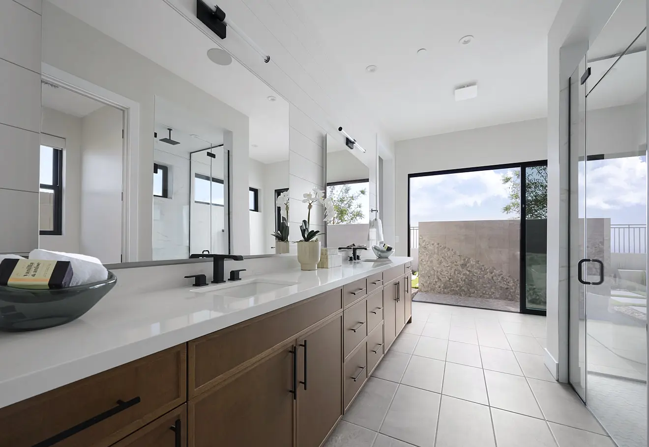 250911_Carlisle_Ridge_P1_009 Modern bathroom in Plan 1 at Carlisle Ridge by Tri Pointe Homes features double sinks, wooden cabinets, large mirrors, black fixtures, and a glass shower. Natural light streams through a big window and glass door, with a potted orchid adding elegance.