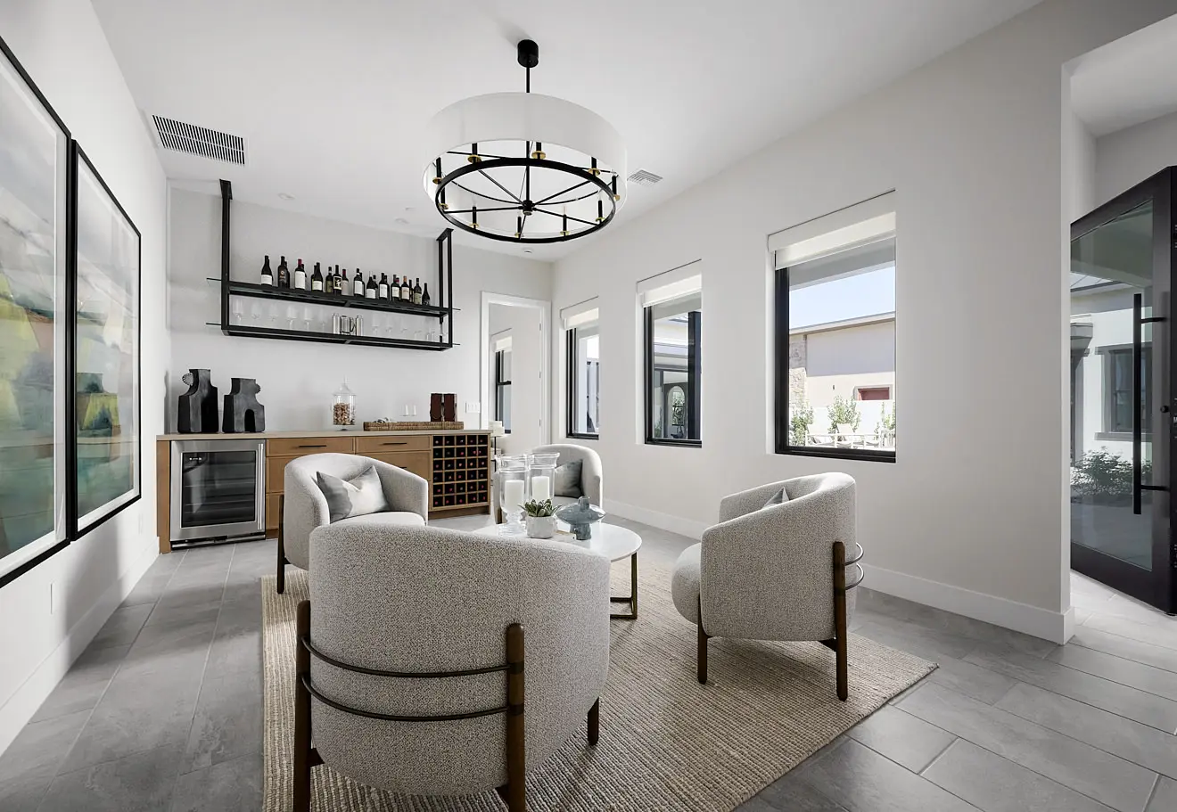 250911_Carlisle_Ridge_P1_011 Modern living room in Plan 1 at Carlisle Ridge by Tri Pointe Homes with four white armchairs around a glass coffee table, a wine bar with shelves and bottles, large wall art, and three windows filling the neutral, contemporary space with natural light.
