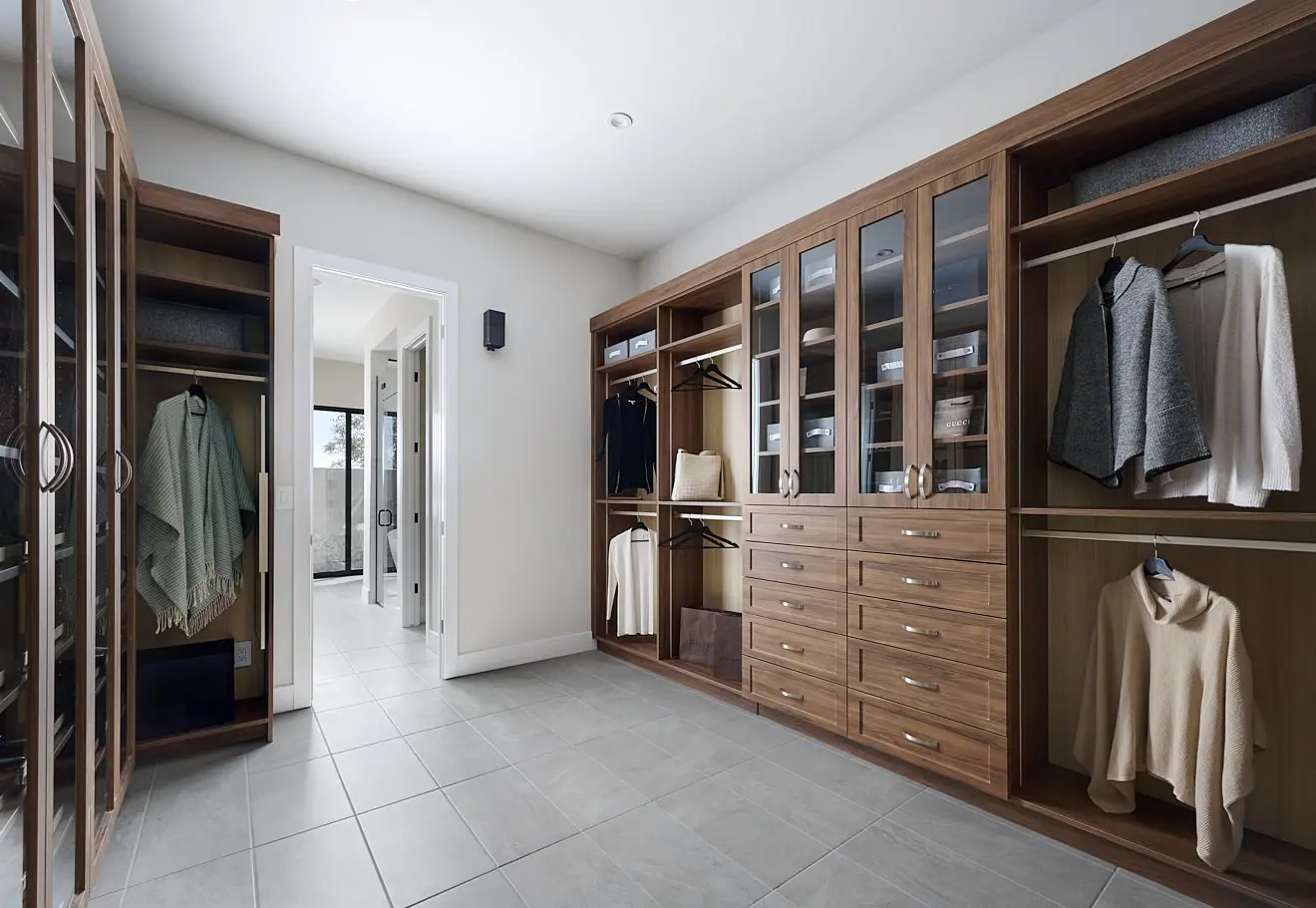 250911_Carlisle_Ridge_P1_013 Modern walk-in closet with wood cabinets, glass doors, open shelves, and drawers in Plan 1 at Carlisle Ridge by Tri Pointe Homes. Clothes and accessories are neatly organized. Light gray tile flooring leads to a bathroom through an open doorway.